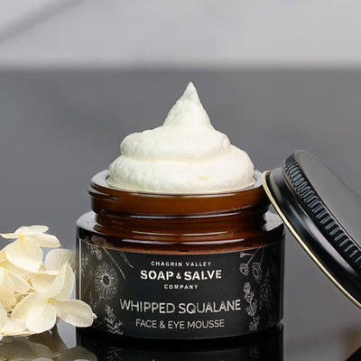 Whipped Squalane Face & Eye Cream in a jar with a creamy top, near white flowers on a reflective surface.