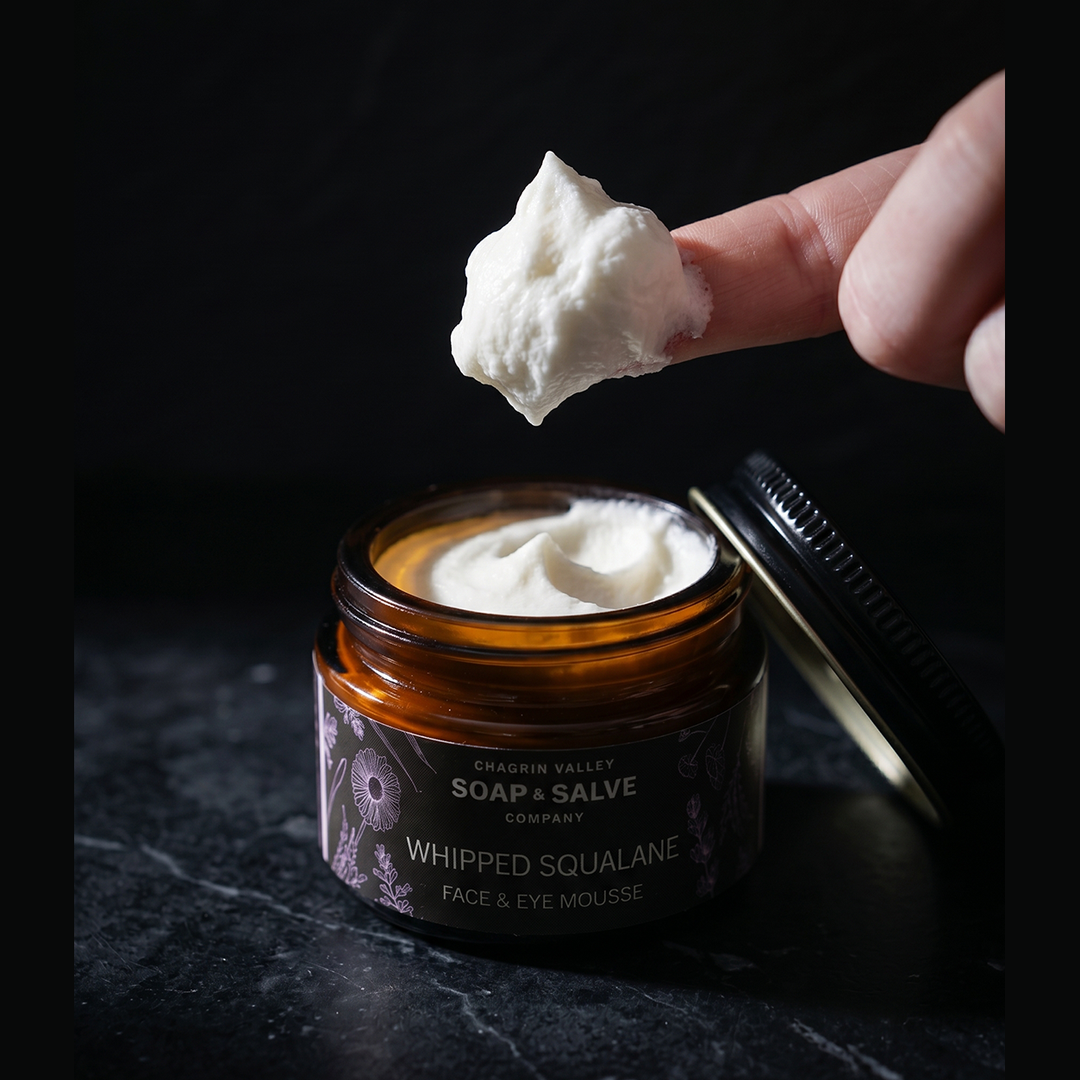 Whipped Squalane Face & Eye Cream in a jar, showing creamy texture on a finger for facial treatment.